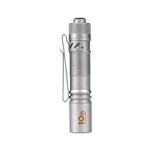 A silver, compact flashlight with a clip and ribbed grip, featuring a "10th Anniversary" label on its side. This titanium EDC flashlight incorporates the Acebeam Pokelit AA Ti (Limited Edition) and NICHIA 519A LED for superior performance.