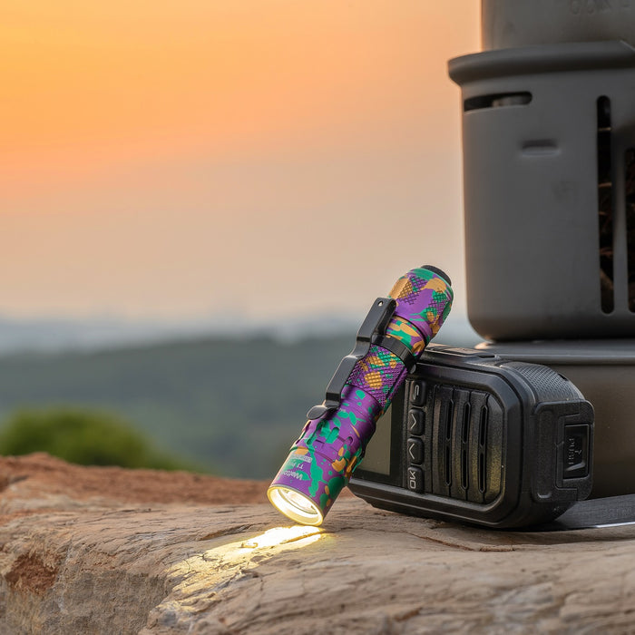 A Weltool T1Pro V2 Colorful Version flashlight stands upright on a rock beside a black device, powered by a 14500 lithium-ion battery, with a blurred sunset and landscape in the background.