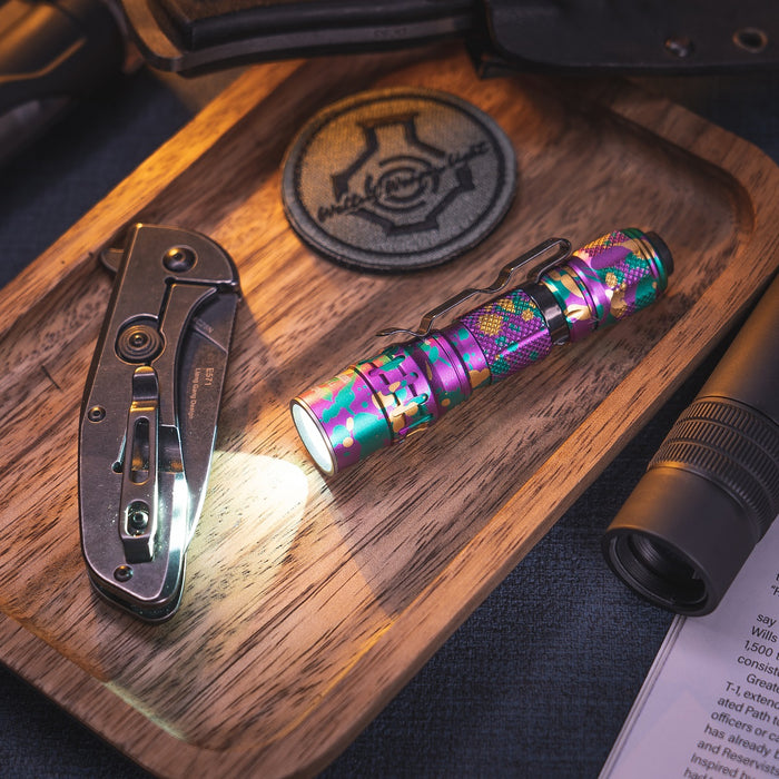 The Weltool T1Pro V2 Colorful Version mini EDC flashlight emits light on a wooden tray next to a folding knife, a decorative patch, and parts of other gadgets.