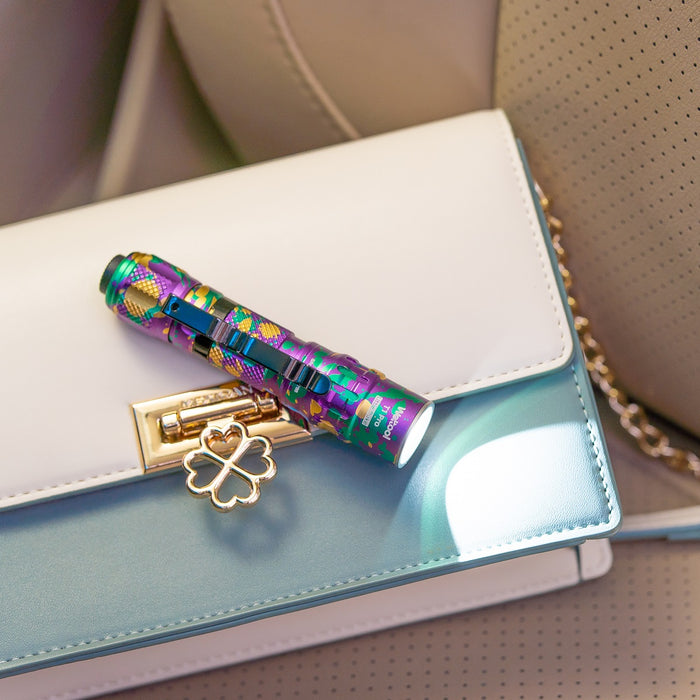 The Weltool T1Pro V2 Colorful Version flashlight rests on a white and teal handbag with a four-leaf clover lock, emitting light onto the bag.