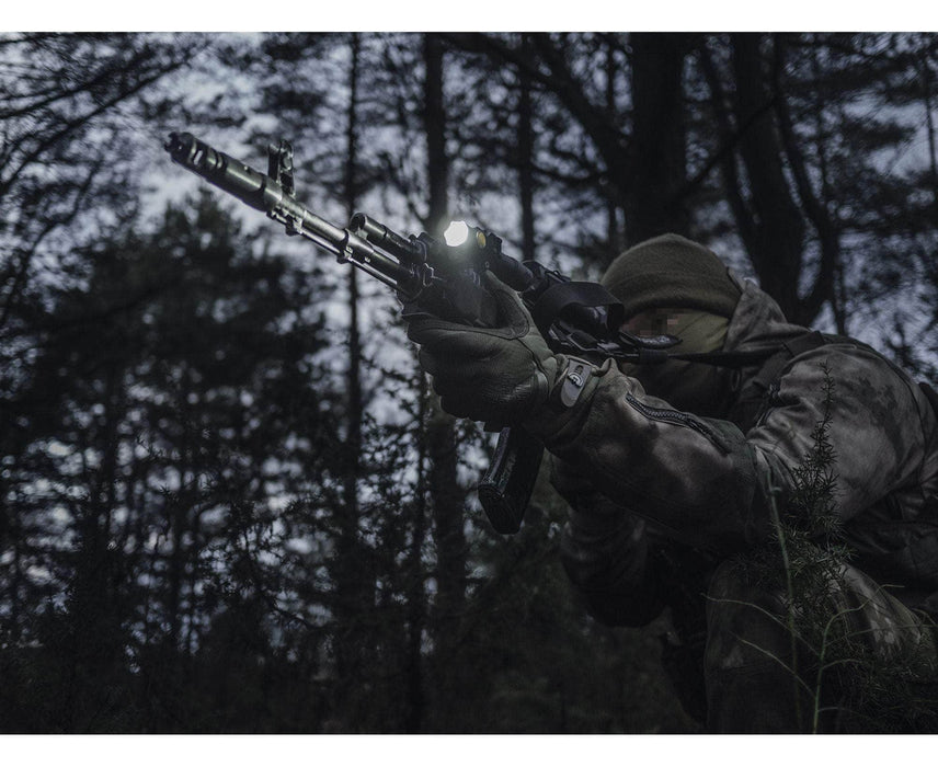 In a dark, forested area, a person in camouflage aims a rifle enhanced with an Armytek Partner C2 Magnet USB - White flashlight.