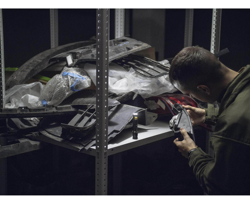 A person examines the Armytek Partner C2 Magnet USB - White car part near a pile of vehicle components on a shelf in a dimly lit space.