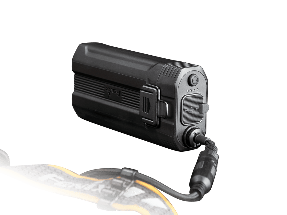 A rectangular black Fenix HP30R V2.0 flashlight with 3000 Lumens illuminates brightly while connected to a charging cable. It includes a small display and buttons on one side, and part of a detachable strap is visible in the foreground.