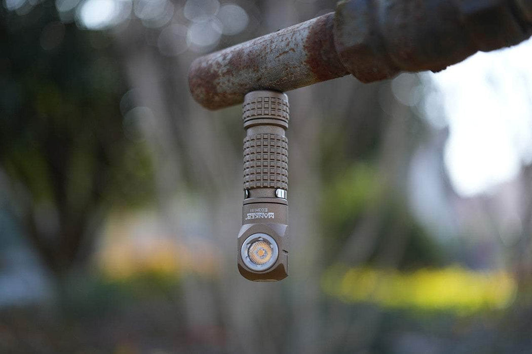 The Manker E03H III, outputting 1200 lumens, magnetically attaches to a rusted pipe outdoors, its textured handle ensuring a firm grip as it points downward.