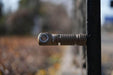 A Manker E03H III flashlight, with 1200 lumens, is magnetically attached to a metal fence outside. Its textured grip provides stability, as the background's blurry colors showcase the vibrant hues of autumn leaves.