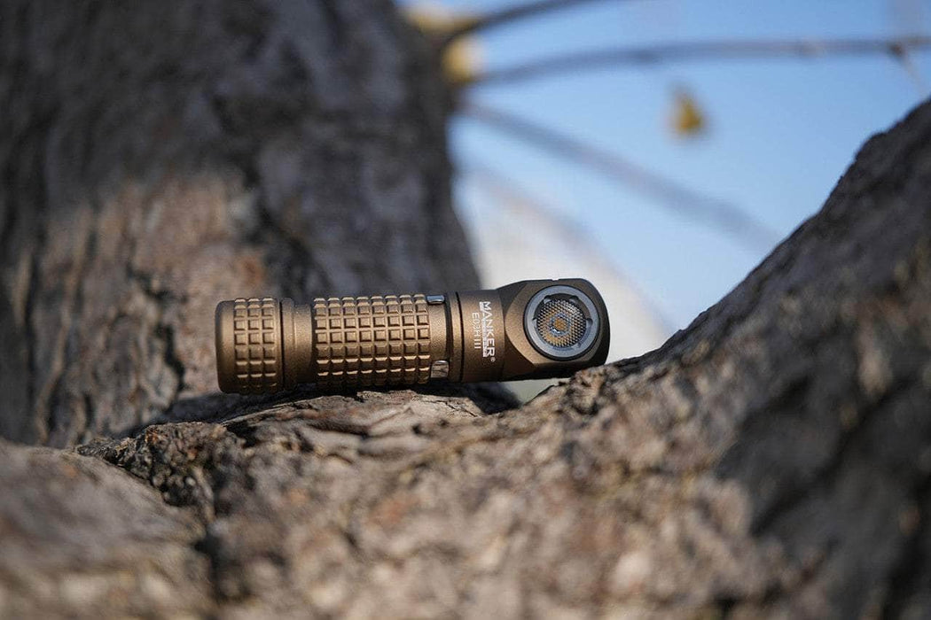 The Manker E03H III flashlight, boasting 1200 lumens, sits on a tree branch against a clear sky ready to outshine any headlamp.