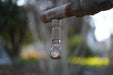 The Manker E03H III flashlight dangles from a weathered metal pipe outdoors, its glow illuminating a softly blurred green and brown background.