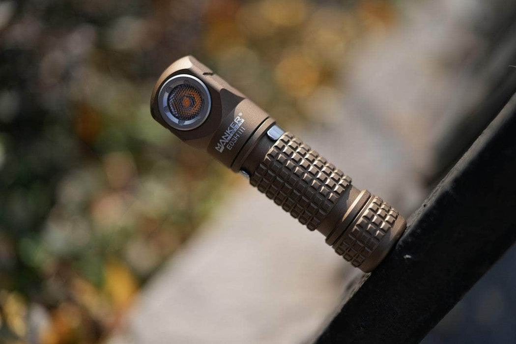 A tan Manker E03H III, a compact everyday carry flashlight with 1200 lumens and a textured grip, rests on a ledge outdoors.