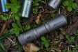 The Manker E05 II - Stonewashed Ti flashlight, boasting 1300 Lumens, rests among green plants, its sturdy design ideal for Everyday Carry, with extra parts nearby ready to assist in any adventure.