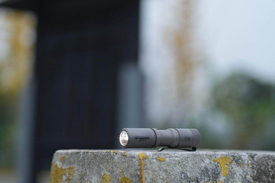 The Manker E05 II - Stonewashed Ti, a sleek titanium flashlight emitting 1300 lumens, sits on a concrete surface with a blurred background emphasizing its everyday carry allure.