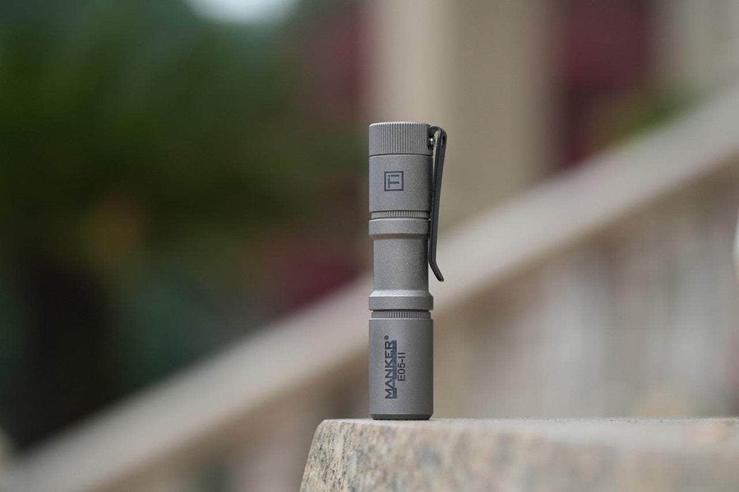 The Manker E05 II - Stonewashed Ti, a compact titanium flashlight with 1300 lumens and a clip, sits outdoors on stone. Its sleek design and powerful brightness make it ideal for everyday carry and essential in any situation.