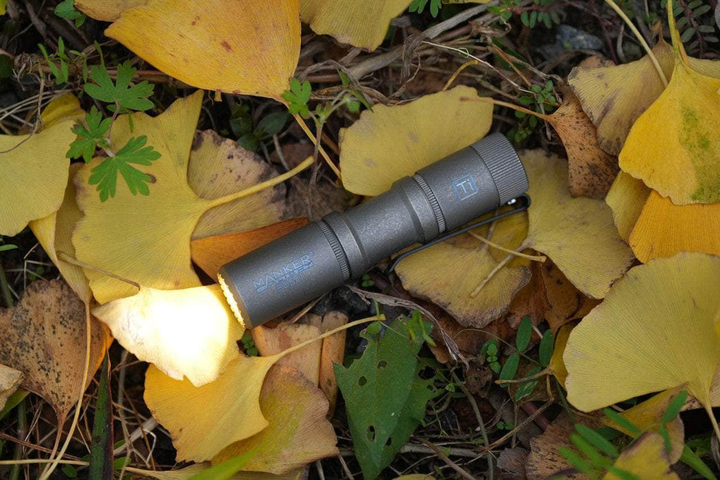 The Manker E05 II - Stonewashed Ti flashlight, emitting 1300 lumens, illuminates vibrant yellow leaves and greenery, making it perfect for everyday carry.