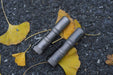 Two Manker E05 II flashlights, crafted from stonewashed titanium, lie on the asphalt amid yellow ginkgo leaves. Ideal for everyday use, these powerful tools deliver 1300 lumens of brightness.