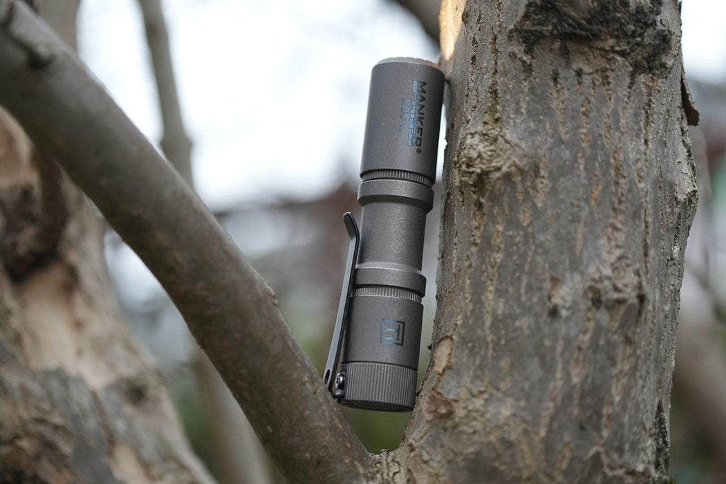 The Manker E05 II - Stonewashed Titanium, a compact flashlight with 1300 lumens, is clipped onto a tree branch and efficiently powered by a 14500 AA battery.