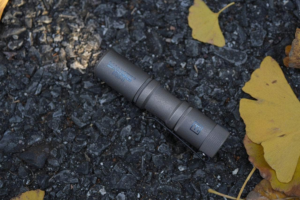 The Manker E05 II Stonewashed Ti flashlight sits on the asphalt among scattered yellow leaves, ideal for everyday carry in gray titanium.