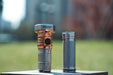 A copper-colored Pocket EDC Flashlight with intricate detailing stands next to a textured, cylindrical metal object, both placed on a flat surface outdoors with a blurred green background. The scene showcases the elegance of the Manker MC13 II Pocket EDC Flashlight - Titanium & Copper's design in its Titanium & Copper blend.