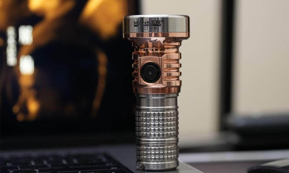 A close-up of a titanium and copper-colored Manker MC13 II Pocket EDC Flashlight - Titanium & Copper standing on a surface, with a blurred background.