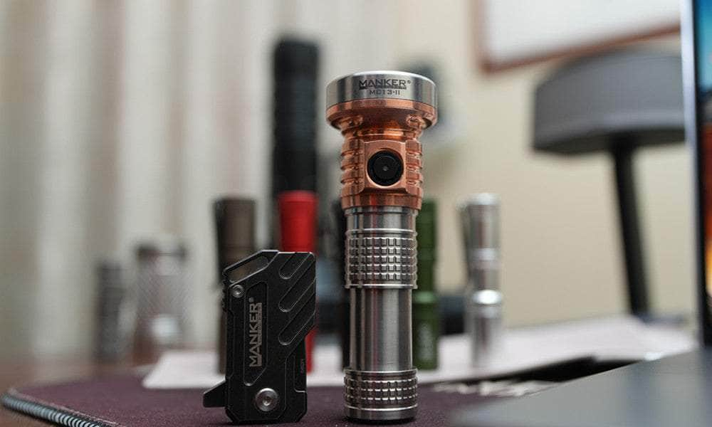 Several Manker flashlights, including the Manker MC13 II Pocket EDC Flashlight - Titanium & Copper, are displayed on a desk with additional flashlights blurred in the background.