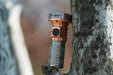 A Manker MC13 II Pocket EDC Flashlight - Titanium & Copper with a sleek metallic body, crafted from Titanium & Copper, is propped against a tree trunk outdoors.