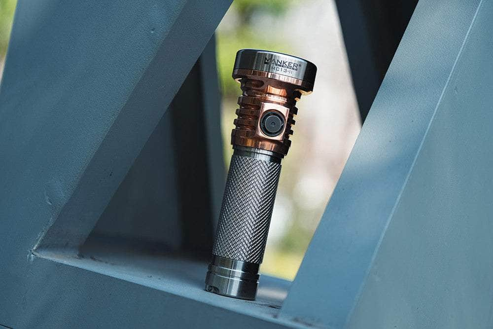 A Manker MC13 II Pocket EDC Flashlight - Titanium & Copper with a textured grip and a sleek titanium & copper finish rests on a geometric structure outdoors.