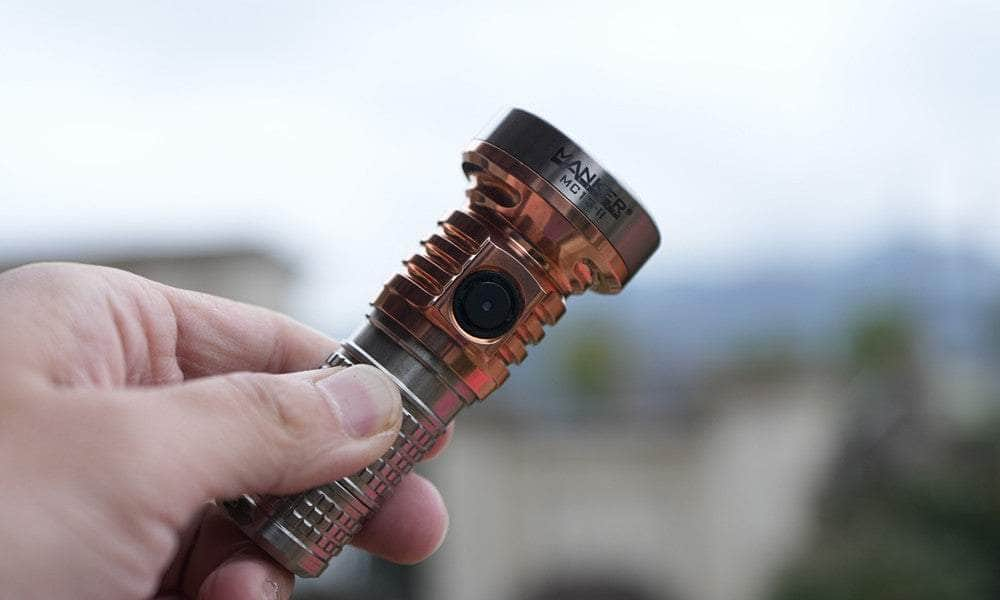 A person holding a Manker MC13 II Pocket EDC Flashlight - Titanium & Copper, metallic cylindrical gadget with a ridged grip and a lens on its front, against an outdoor blurred background.