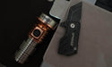 A compact Manker MC13 II Pocket EDC Flashlight - Titanium & Copper and a foldable knife, both marked with the brand "Manker," are placed on a light brown surface next to a silver and black electronic device.