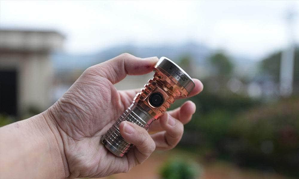 A hand is holding a small, metallic Manker MC13 II Pocket EDC Flashlight - Titanium & Copper with a prominent button on its side, against an outdoor background.