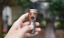 A hand holding a metallic Pocket EDC Flashlight with the brand name "Manker" and model "MC13 II Pocket EDC Flashlight - Titanium & Copper" engraved on top, set against a blurred outdoor background.