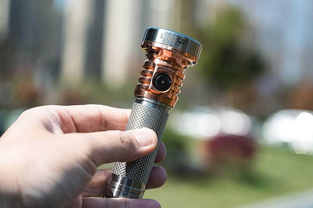 A hand holding a Manker MC13 II Pocket EDC Flashlight - Titanium & Copper with a textured grip and a lens-like feature on the head, set against a blurred outdoor background.