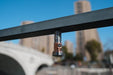 A Manker MC13 II Pocket EDC Flashlight - Titanium & Copper is magnetically attached to a metal rail, with blurred buildings and a bridge in the background.