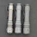 Three metallic flashlights with belt clips arranged in a row against a dark background. Each flashlight, including the ReyLight LANapple - Ti, boasts slightly different shades and finishes. One features the luminous Titanium Nichia 519a LED, while another is powered by a rechargeable 14500 battery.