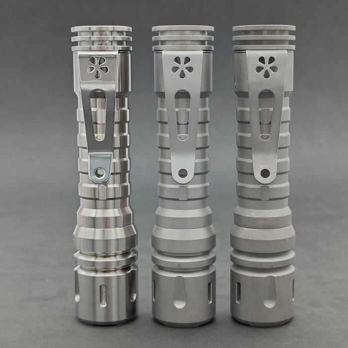 Three ReyLight LANapple - Ti flashlights with decorative vent holes are standing upright on a gray surface. These silver titanium flashlights feature ridged bodies, sturdy clips for attachment, and are compatible with rechargeable 14500 batteries.