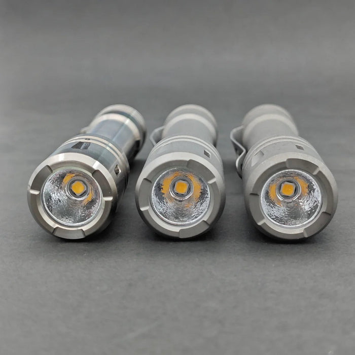 Three ReyLight LANapple - Ti flashlights are aligned side by side, each with a titanium gray body and an LED bulb visible at the front. The background is a plain, dark surface. These sleek tools run on rechargeable 14500 batteries for extended use.