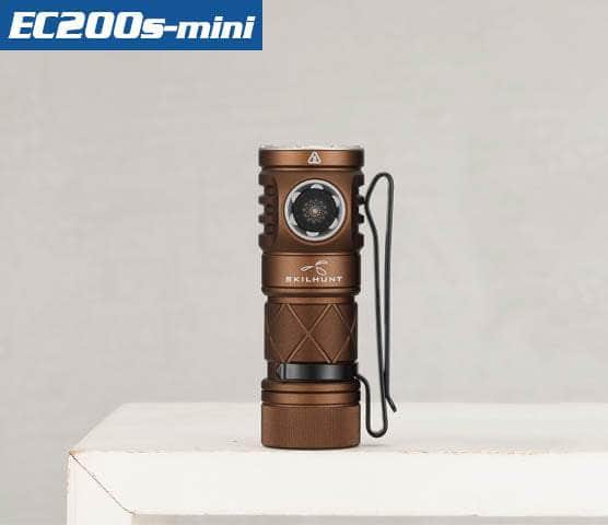 A brown Skilhunt EC200S-Mini EDC flashlight is standing upright on a white surface, with a pocket clip attached to its side. The product name is displayed in the top left corner. It features a USB-C charging port for convenience.