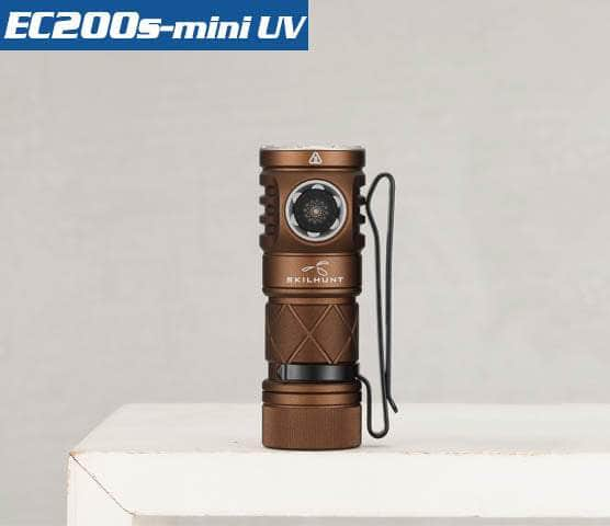 A compact, bronze-coloured Skilhunt EC200S-Mini UV with a black clip, capable of emitting a 2100-lumen cool white beam or 365nm UV beam, displayed vertically on a white surface with a plain grey background.