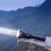 The Weltool F11R, a rechargeable long-range LED flashlight that emits a strong beam of light, sits on a rock ledge with misty, tree-covered mountains in the background.