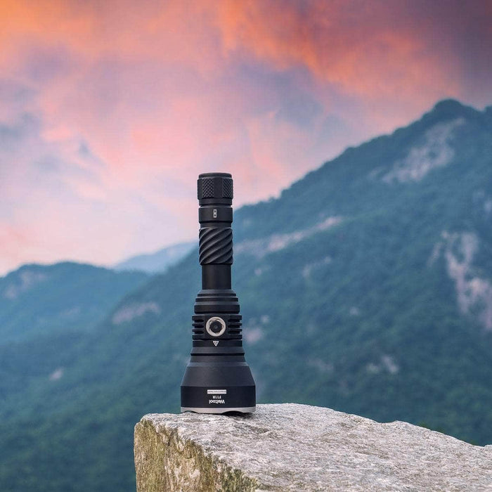 The Weltool F11R, a rechargeable long-range LED flashlight with a USB-C charging structure, stands on a rock, silhouetted against mountains and a dramatic sky in the background.