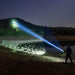 A person shines a Weltool F11R rechargeable long-range LED flashlight across a dark landscape, illuminating trees on a hill in the distance.