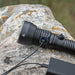 The Weltool F6R, a high-lumen rechargeable black flashlight, is shown on a rock surface, being charged via a USB cable connected to a power source.