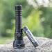 A high-lumen Weltool F6R rechargeable flashlight stands upright on a rock, next to a 15000mAh battery labeled "Weltool XR 35L 3.6V". The background is blurred greenery.