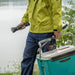 Person in a green jacket holding a Weltool F6R, fishing rod, and a blue cooler near a lake.