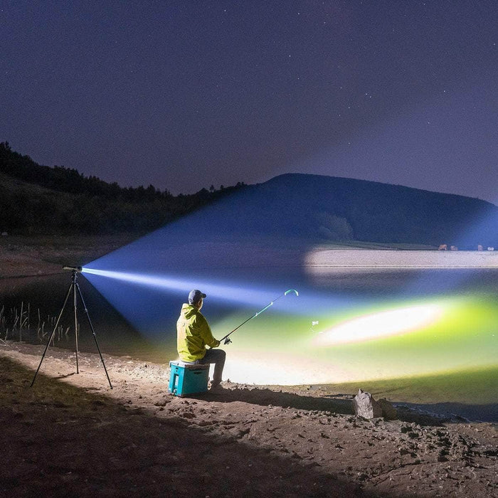 A person in a yellow jacket sits on a green cooler, fishing at night by a lake. The surroundings are illuminated by the high-lumen Weltool F6R rechargeable flashlight mounted on a tripod.