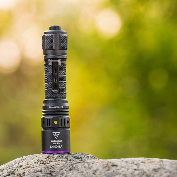 The Weltool M4-365 UV flashlight, emitting a glow at 365nm, rests on a rock against a blurred green and yellow background.