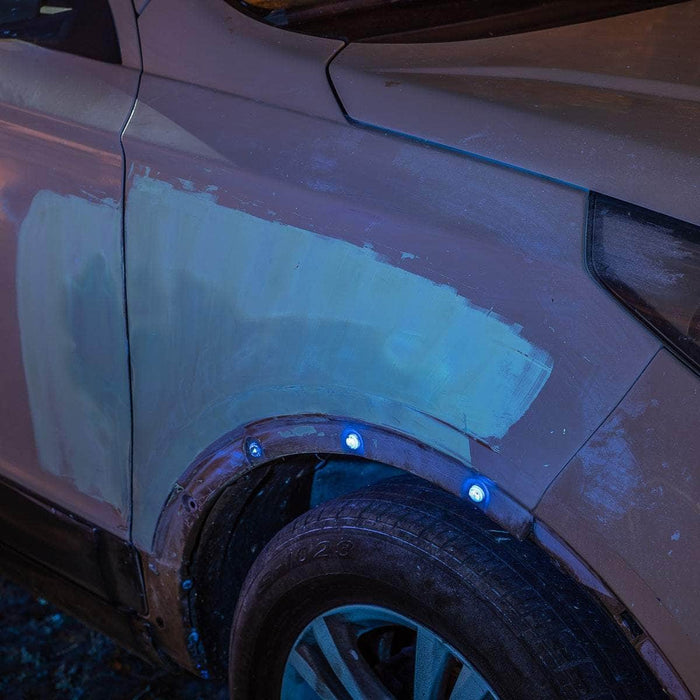 Car with a partially repaired front fender, showing unpainted patches and visible attachment screws around the wheel arch. Under the illumination of a Weltool M4-365 UV flashlight, the imperfections are even more pronounced, highlighting areas in need of attention.