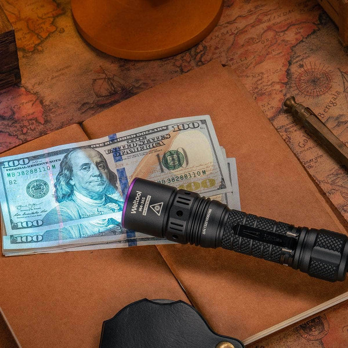 The Weltool M4-365 UV flashlight, emitting a powerful radiation flux of 365nm, unveils the security features on a stack of hundred-dollar bills placed on an old map-themed desk surface.