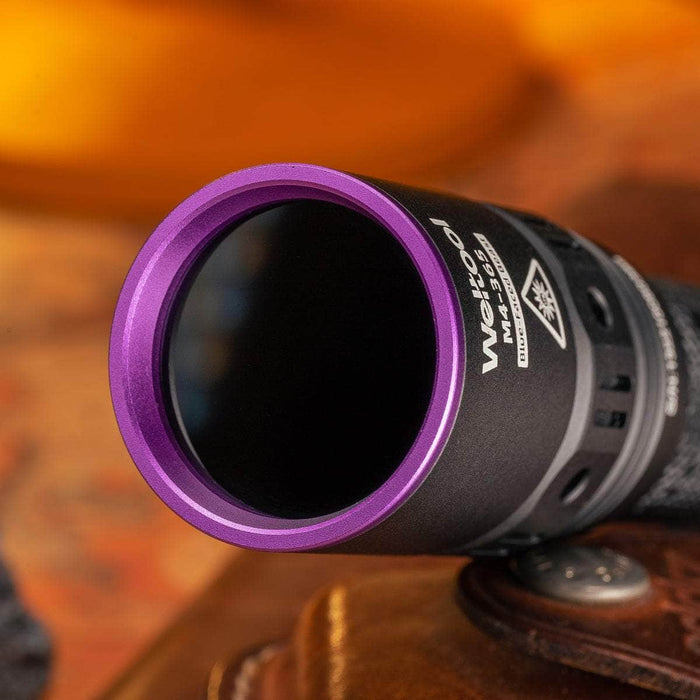 Close-up of the Weltool M4-365 UV flashlight in black metal, showcasing a purple-rimmed lens and labeled with "WO" and "MA-53A," designed for 365nm high radiation flux. The background is blurred with warm tones.