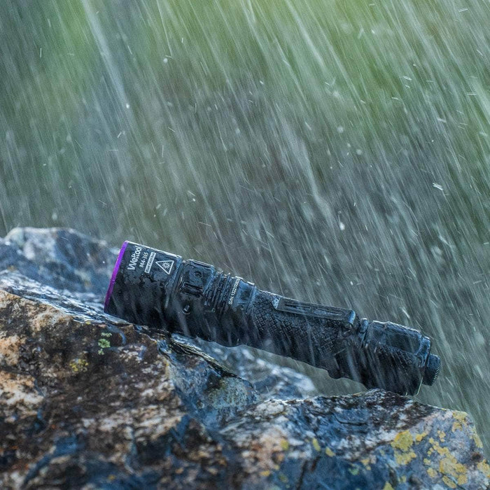 The Weltool M4-365 UV flashlight rests on a wet rock in the rain, emitting 365nm wavelength light as water droplets cascade around it.