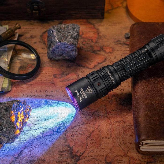 Using the Weltool M4-365 UV flashlight, which emits 365nm blue light, a vintage map is illuminated. Nearby, there are a magnifying glass, stones, and a leather-bound book.