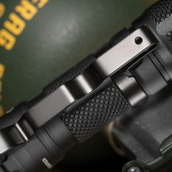 Close-up of the black textured grip of the Weltool T1 Pro V2 EDC LED flashlight, featuring a metallic clip, set against a blurred background with yellow text and green colors—ideal for emergency preparedness.