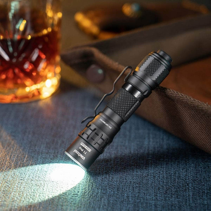 The Weltool T1 Pro V2, a compact black flashlight, stands on its end, casting a beam of light onto a blue surface with a glass of whiskey and a brown bag in the background. Ideal for everyday carry, it efficiently uses a 14500 lithium-ion battery.